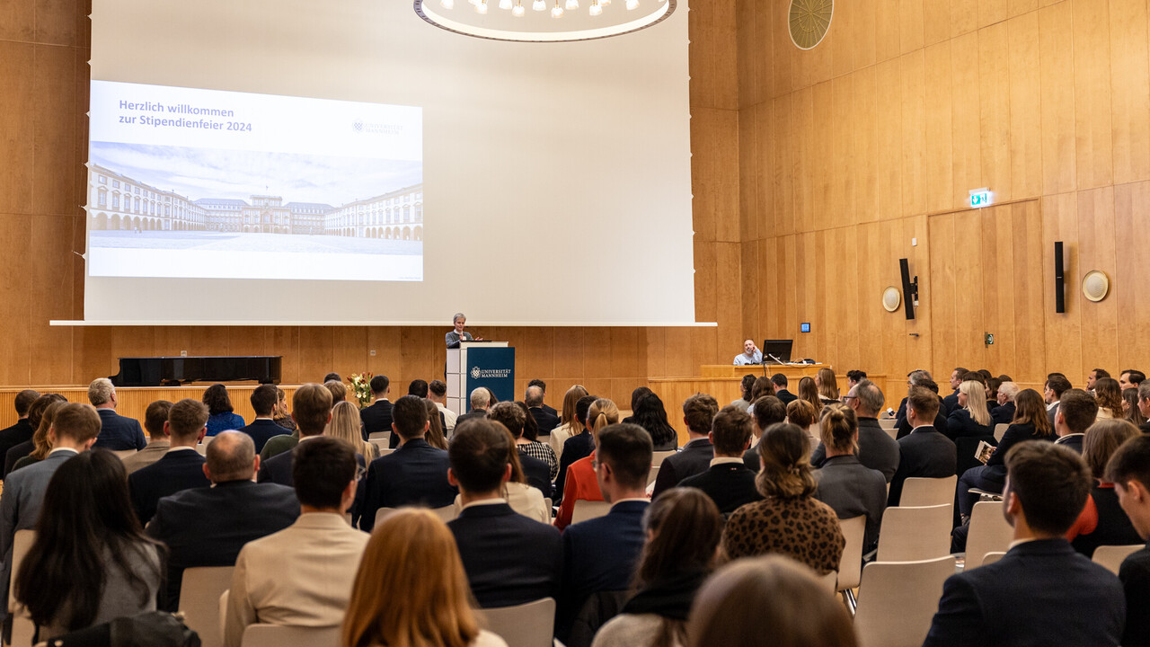 Dutzende Menschen sitze in einem festlich beleuchteten Saal und schauen auf die Bühne, auf der eine Person steht und spricht.