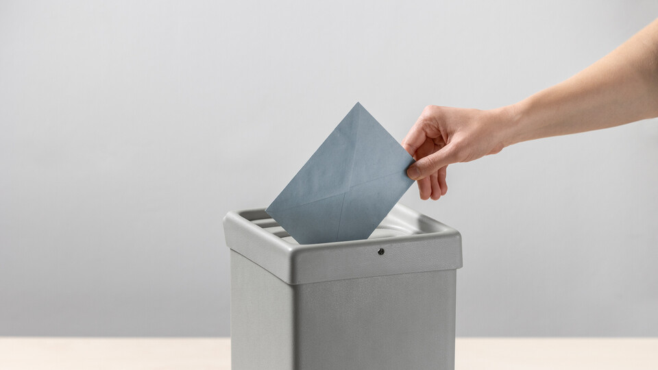 Eine Person steckt einen blauen Umschlag in eine Wahlurne.