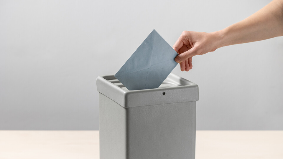 Eine Person steckt einen blauen Umschlag in eine Wahlurne.