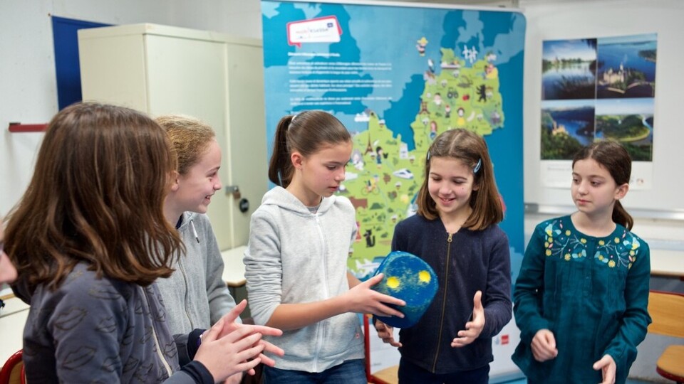 Fünf Kinder reichen sich gegenseitig einen blauen Würfel. Im Hintergrund, eine Karte von Frankreich und Deutschland.