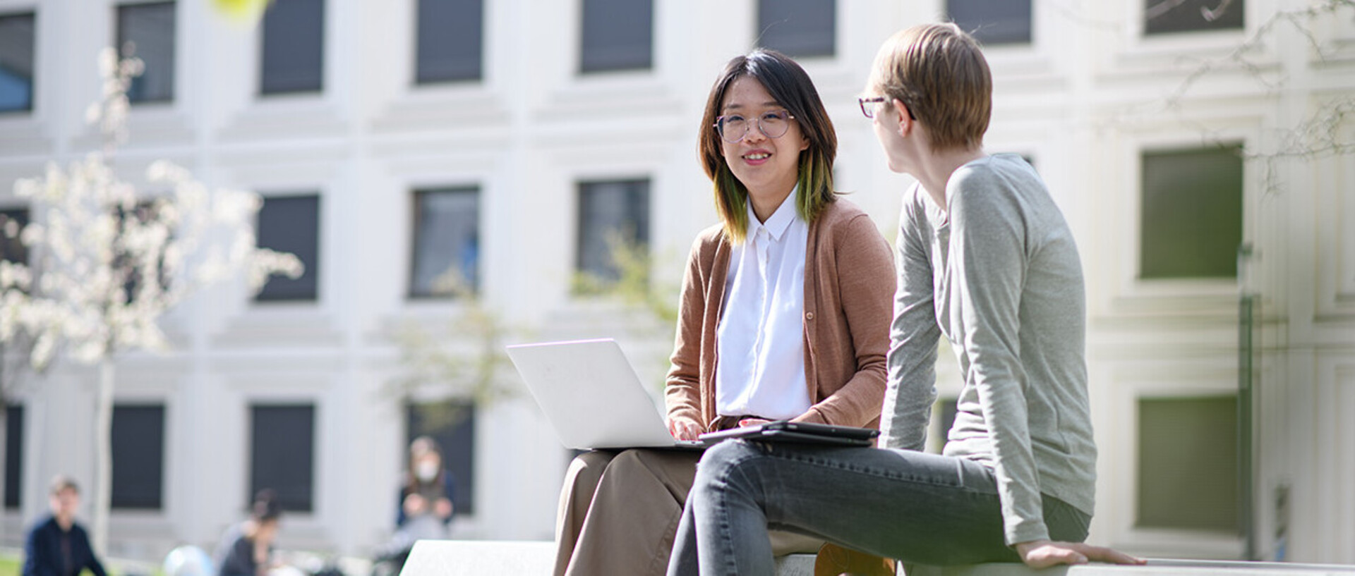 Two women are sitting on a bank in the green courtyard of the B6 building and are looking with a smile at a laptop screen.