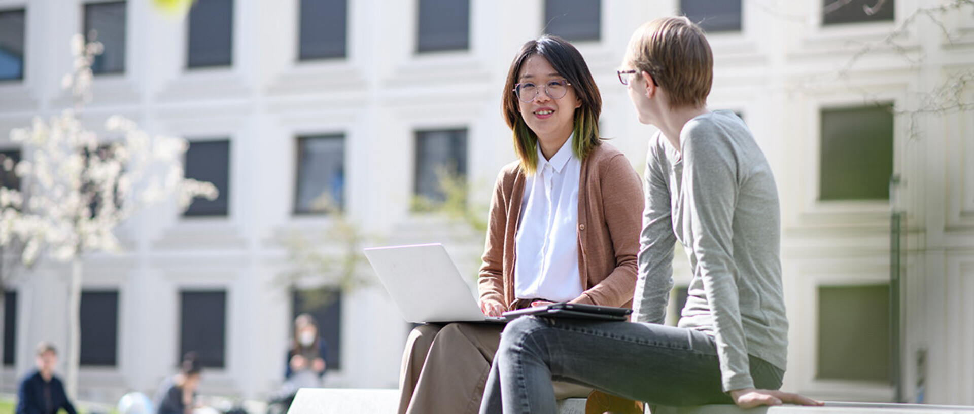 Two women are sitting on a bank in the green courtyard of the B6 building and are looking with a smile at a laptop screen. 