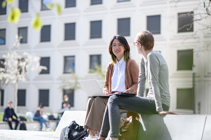 Two women are sitting on a bank in the green courtyard of the B6 building and are looking with a smile at a laptop screen. 