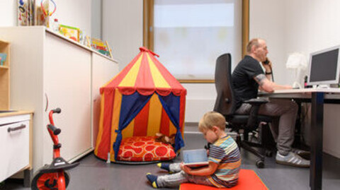 Ein Mitarbeiter sitzt am Schreibtisch im Eltern-Kind-Zimmer und telefoniert. Links neben ihm steht ein Kinderspielzelt und auf einer Matratze sitz ein kleiner Junge und liest ein Buch.