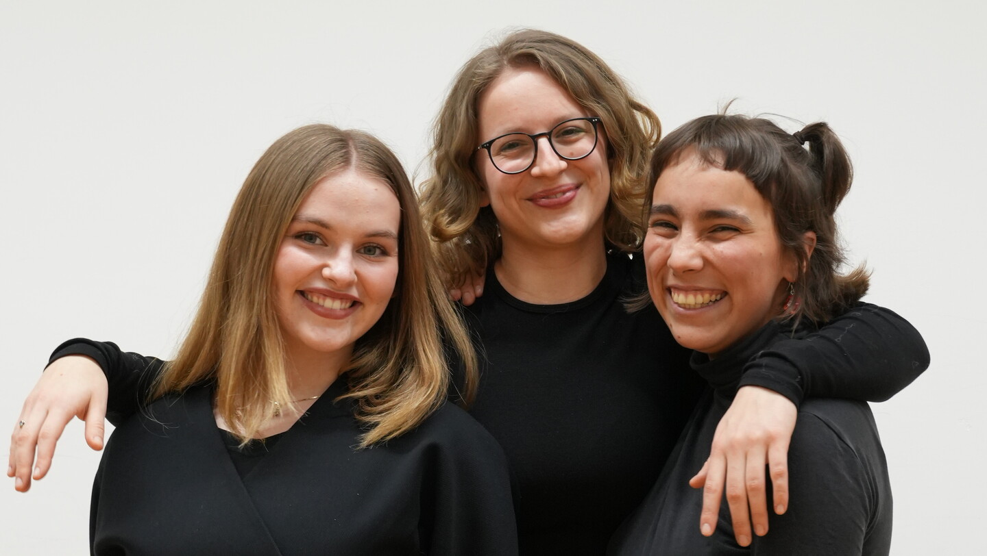 Drei schwarzgekleidete Personen stehen nebeneinander vor einer weißen Wand.