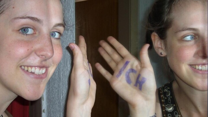 Eine junge Frau steht vor dem Spiegel. Im Spiegel erkennt man den Schriftzug "Ich" auf der Innenseite ihrer Handfläche.