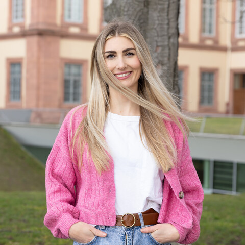Eine lächelnde Person trägt ein weißes T-Shirt, sowie eine blaue Jeans und einen pink-farbenen Cardigan. Die Person steht auf einer Wiese vor dem Schloss der Uni Mannheim und heißt Hannah Müller.