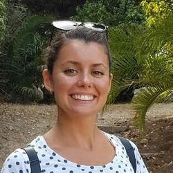 Nikol Toteva hat dunkelbraune Haare, welche sie in einem Zopf trägt. Sie trägt eine Sonnenbrille, einen weißes T-Shirt mit dunkelblauen Punkten und eine Rucksack. Im Hintergrund sind Palmen zu sehen.