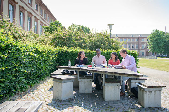 Zwei Frauen und zwei Männer sitzen neben der Mensawiese an einem Tisch und arbeiten fröhlich zusammen. Vor ihnen auf dem Tisch stehen Getränke und Bücher.