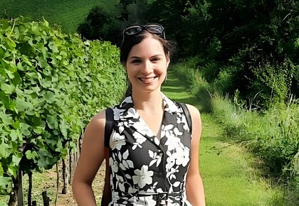 Andreea Iana steht im Sonnenschein auf einem Weinberg. Sie trägt ein geblümtes schwarz-weißes Kleid und eine Sonnenbrille auf dem Kopf.