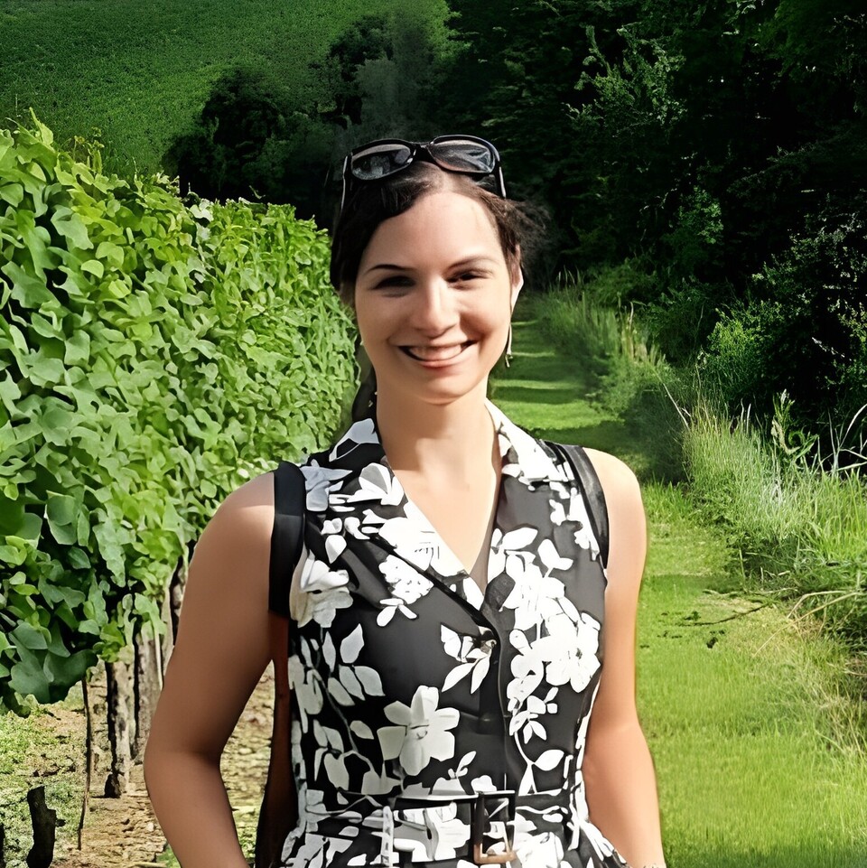 Andreea Iana steht im Sonnenschein auf einem Weinberg. Sie trägt ein geblümtes schwarz-weißes Kleid und eine Sonnenbrille auf dem Kopf.