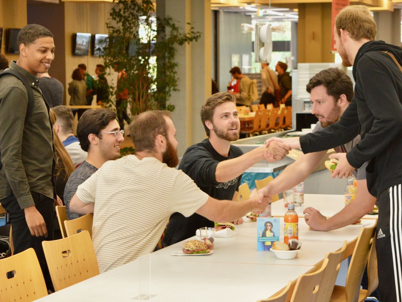 Sechs Studenten sitzen in der Mensa an einem Tisch. Sie verabschieden sich mit einem Handschlag.