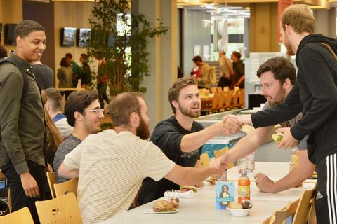 Sechs Studenten sitzen in der Mensa an einem Tisch. Sie verabschieden sich mit einem Handschlag.