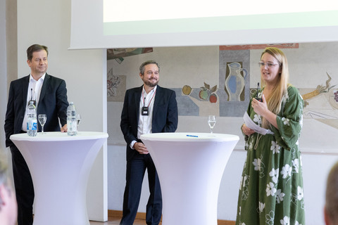 Drei Personen in schicker Kleidung stehen nebeneinander hinter zwei Stehtischen und reden.