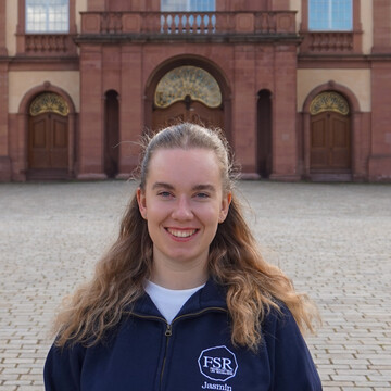 Jasmin trägt einen dunkelblauen Pullover der Fachschaft, hat lange blonde Haare und lächelt freundlich in die Kamera. Im Hintergrund ist der Mittelbau des Mannheimer Schlosses