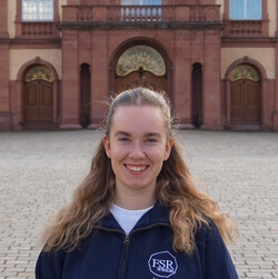 Jasmin is wearing a dark blue pullover of the departmental student committee. She has long, blond hair and is smiling at the camera. In the background, the Mittelbau of the Schloss can be seen.