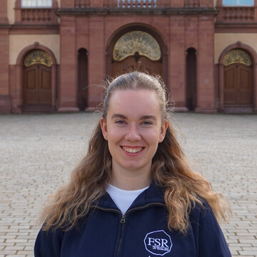 Jasmin is wearing a dark blue pullover of the departmental student committee. She has long, blond hair and is smiling at the camera. In the background, the Mittelbau of the Schloss can be seen.