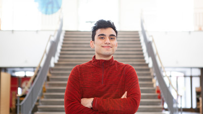 Eine Person trägt einen rot-schwarz mellierten Pullover und steht in einem Gebäude vor einer Treppe. Die Person heißt Hyder Amin. 