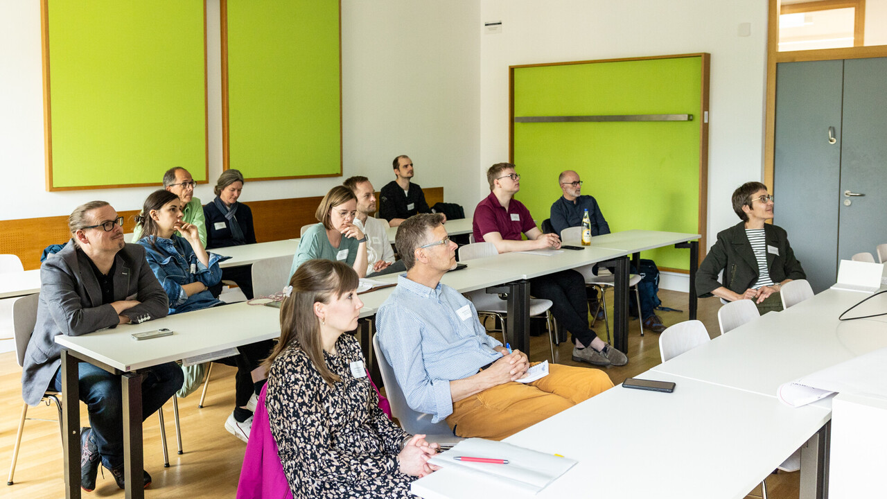 Teilnehmer*innen eines Workshops sitzen in mehreren Reihen an Tischen in einem hellen und freundlichen Seminarraum und hören aufmerksam zu