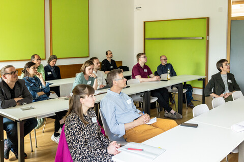 Teilnehmer*innen eines Workshops sitzen in mehreren Reihen an Tischen in einem hellen und freundlichen Seminarraum und hören aufmerksam zu