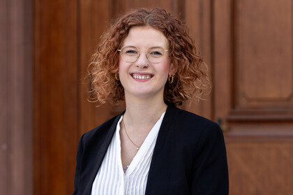 Larissa Will steht vor dem Haupteingang des Schlosses im Ehrenhof. Sie hat Locken, trägt eine Brille und einen schwarzen Blazer. 