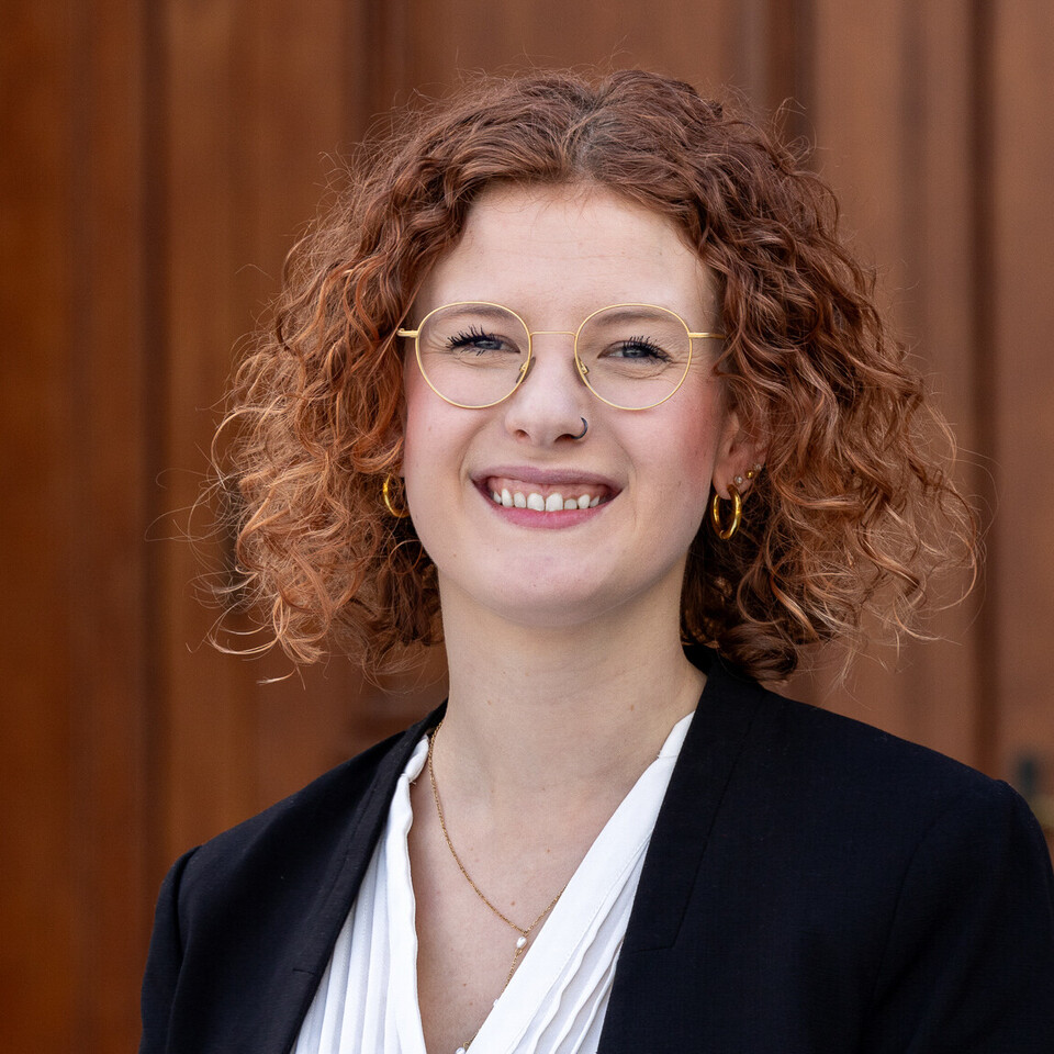 Larissa Will steht vor dem Haupteingang des Schlosses im Ehrenhof. Sie hat Locken, trägt eine Brille und einen schwarzen Blazer. 
