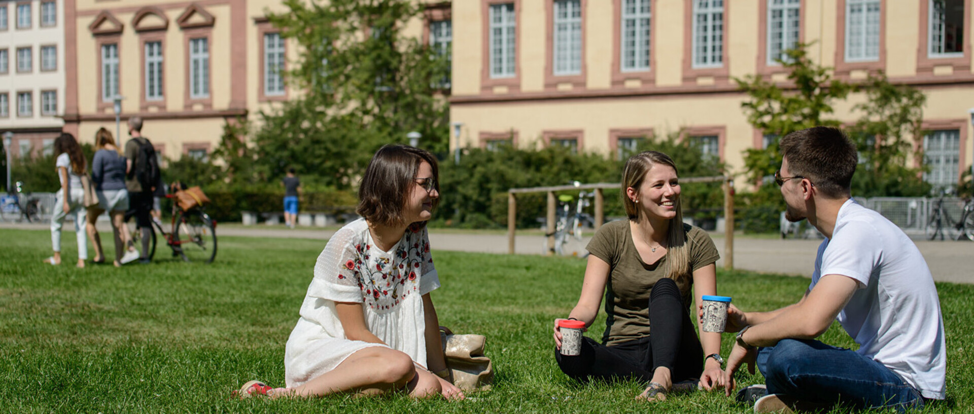 Zwei Studentinnen und ein Student sitzen auf der Mensawiese. Sie tragen sommerliche Kleidung und halten To-Go-Becher in ihren Händen.
