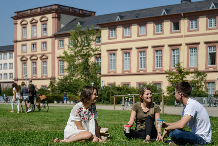 Zwei Studentinnen und ein Student sitzen auf der Mensawiese. Sie tragen sommerliche Kleidung und halten To-Go-Becher in ihren Händen.
