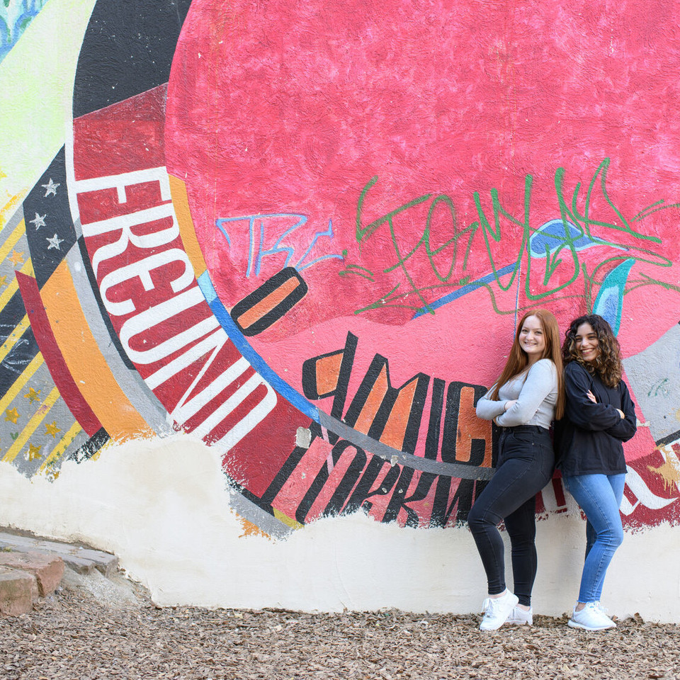 Two students stand in front of a wall with graffitis and laugh.