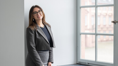 Porträtbild von Joana Furhmann. Sie hat halblange braune Haare und eine Brille, trägt einen grauen Blazer und einen grauen Rock und steht neben einem Fenster.