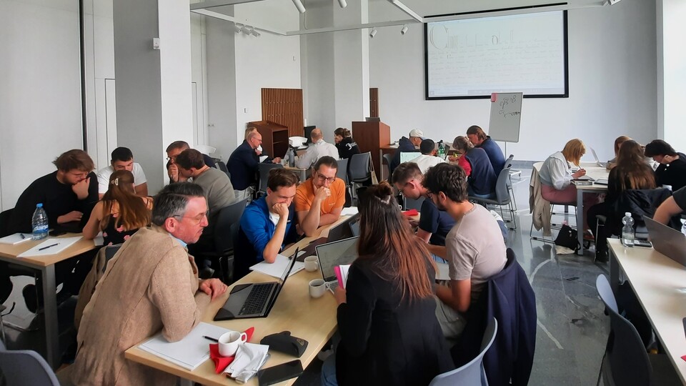 Die Teilnehmenden der Summer School diskutieren in Kleingruppen an Tischen in einem hellen Konferenzraum. Laptops sind aufgeklappt, und eine Präsentation wird an die Wand projiziert.