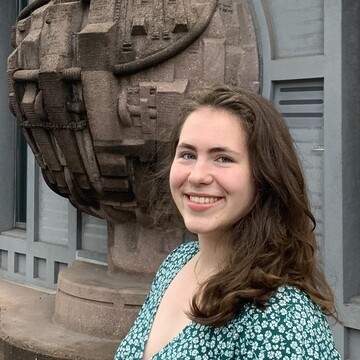Maja Schwerdtle mit schulterlangen braunen Haaren trägt eine hellgrüne Bluse mit Blumenmuster und lächelt freundlich in die Kamera mit dem Gebäude A1 der Universität im Hintergrund.