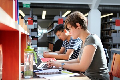 Eine Studentin und ein Student sitzen in der Bibliothek nebeneinander und lernen.