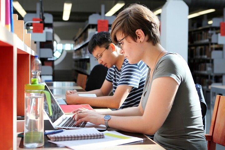 Eine Studentin und ein Student sitzen in der Bibliothek nebeneinander und lernen.