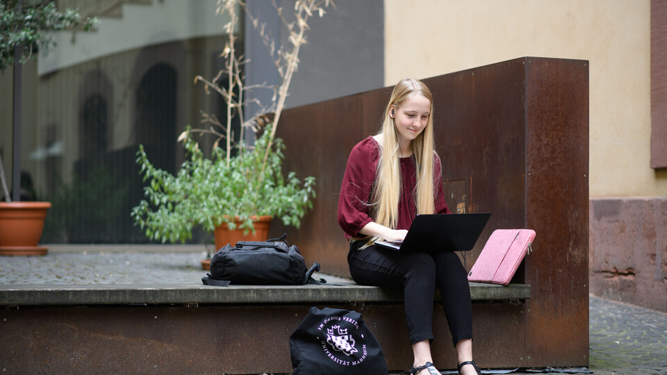 Eine Studentin sitzt mit ihrem Laptop auf dem Schoß draußen und hat Kopfhörer in den Ohren.
