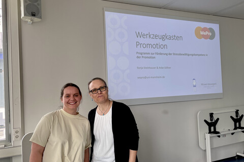 2 weibliche Personen stehen in einem Seminarraum vor einer Powerpointpräsentation auf dem Beamer zum Thema "Werkzeugkasten Promotion"