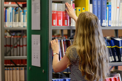 Eine Studentin steht vor einem Bücherregal in der Bibliothek und ist dabei ein Buch herauszuziehen. Sie trägt welliges, blondes Haar und ein schwarz-weiß gestreiftes T-Shirt.