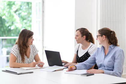 Drei Personen sitzen am Tisch eines Seminarraums und unterhalten sich.