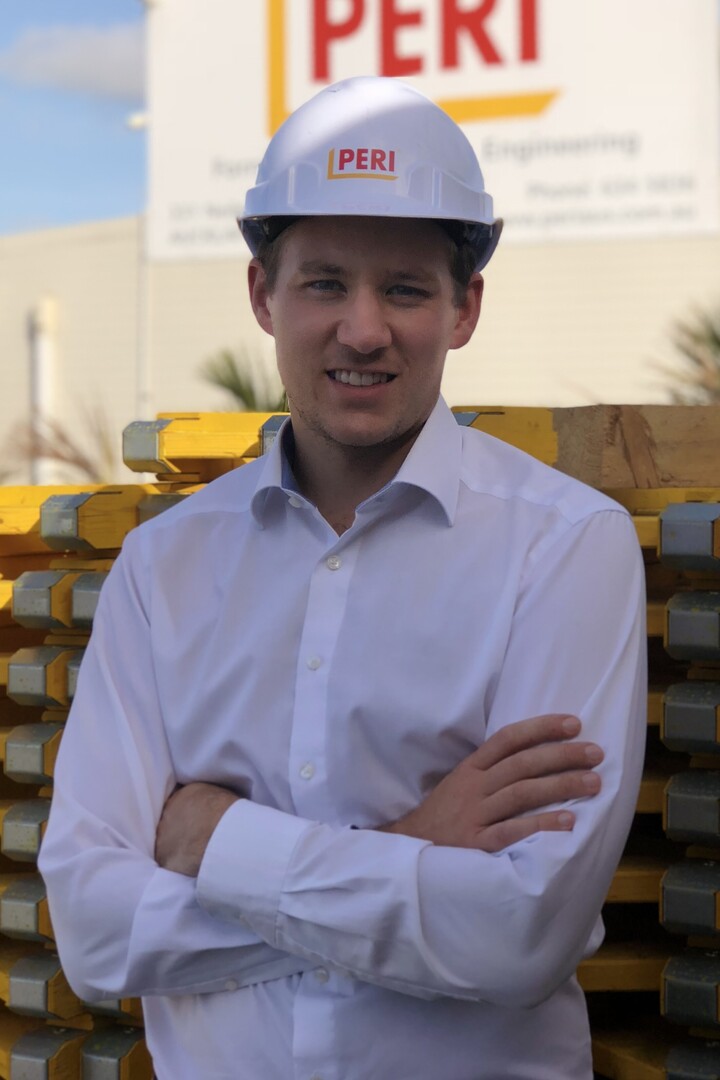 Markus Poeplau steht in weißem Hemd und mit weißem Bauhelm mit PERI-Schriftzug mit verschränkten Armen vor einer Fabrikhalle mit großem PERI-Schild 