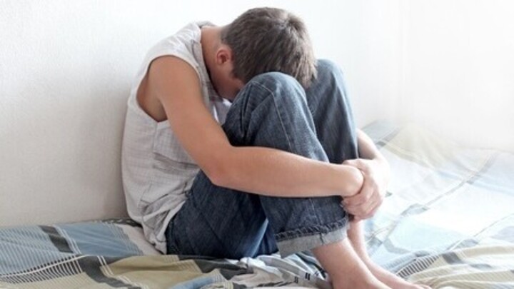 Symbolbild: Ein Jugendlicher sitzt auf einem Bett an der Wand gelehnt und vergräbt seinen Kopf zwischen seinen angewinkelten Beinen