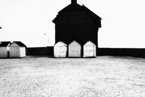 Schwarz-weiß Foto: Vor einem großen schwarzen Haus sind drei kleine Hütten, links stehen zwei weitere Hütten.