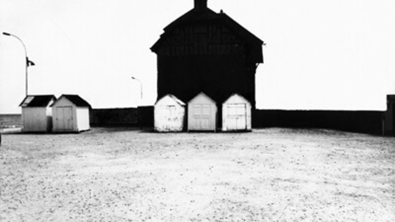 Schwarz-weiß Foto: Vor einem großen schwarzen Haus sind drei kleine Hütten, links stehen zwei weitere Hütten.