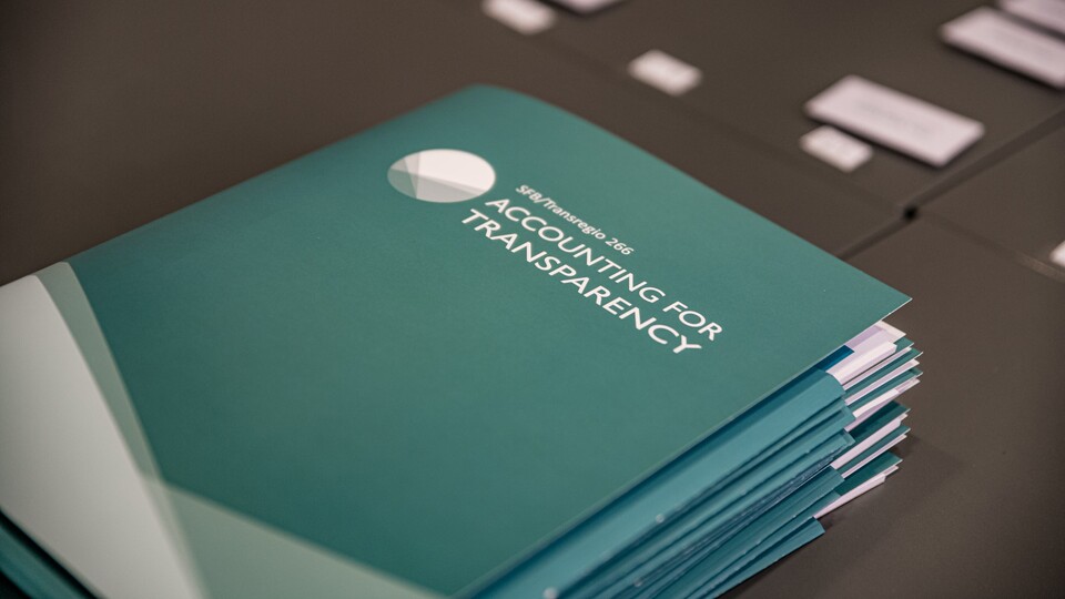 Türkisfarbene Mappen liegen auf einem schwarzen Tisch. Die Mappe haben die weiße Aufschrift "SFB/Transregio 266, Accounting for Transparency".