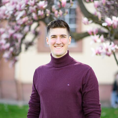 Eine lächelnde Person trägt einen weinroten Pullover sowie eine schwarze Hose und steht auf einer Wiese vor einem Magnolienbaum. Die Person heißt Jonas Wenzel.