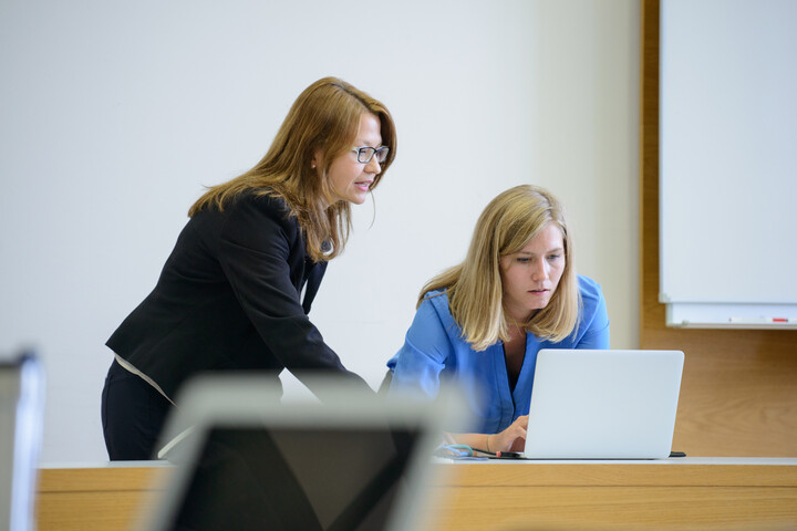 Zwei Personen stehen im Hörsaal, eine beugt sich über einen Laptop, die andere erklärt etwas darauf.