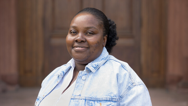 Henrietta Oppong-Adjei. She is wearing a blue jeans jacket over a white shirt and is standing in front of brown door.