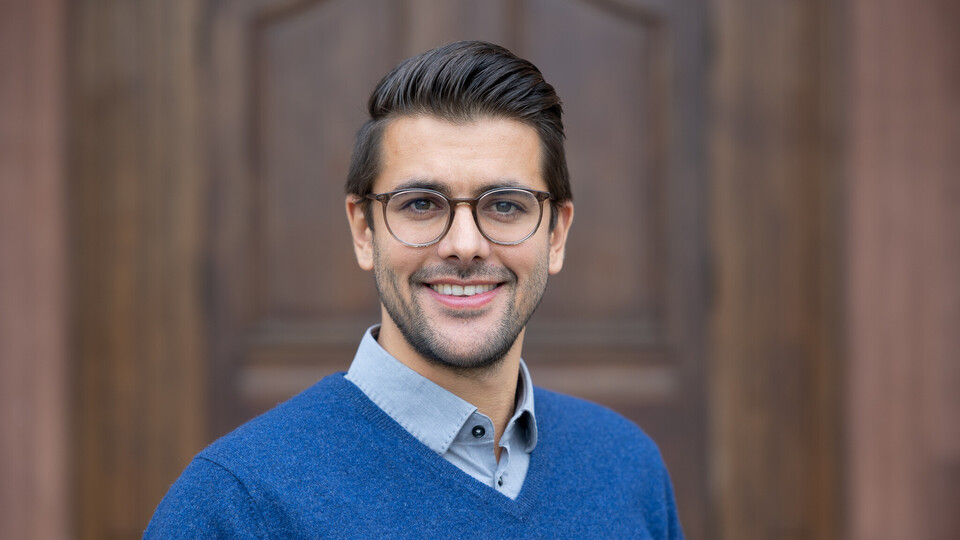 Lukas Hack. He is wearing a blue sweater and is standing in front of a brown door.