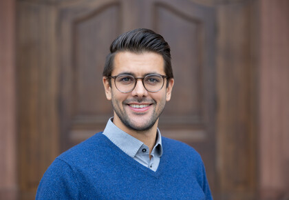 Lukas Hack. He is wearing a blue sweater and is standing in front of a brown door.