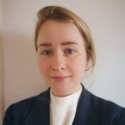 Eine Person trägt ein weißes Oberteil sowie einen dunkelblauen Blazer und steht vor einer weißen Wand. Die Person heißt Marie Barrera. 