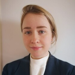 Eine Person trägt ein weißes Oberteil sowie einen dunkelblauen Blazer und steht vor einer weißen Wand. Die Person heißt Marie Barrera.